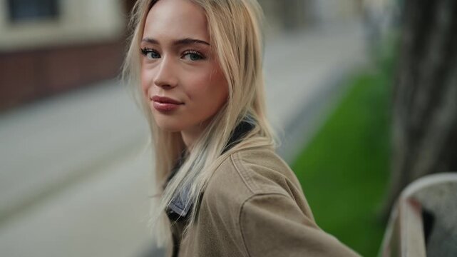 Gorgeous young blonde woman with natural makeup looking over her shoulder and smiling flirtatiously at the camera while standing on a city street in slow motion on a beautiful day
