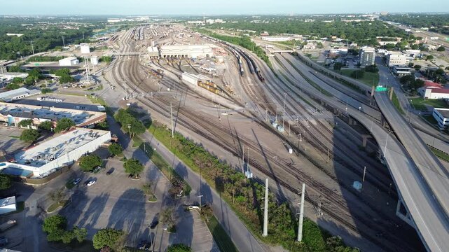 Extensive railroad networks branch through sprawling industrial landscape alongside interstate overpasses. Long, dramatic shadows stretch across grassy patches near retail sites, Fort Worth, TX