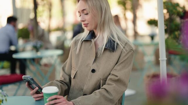 Smiling blonde woman sitting at an outdoor cafe table, enjoying a cup of coffee while browsing social media on her smartphone and smiling pleasantly in a sunny urban setting