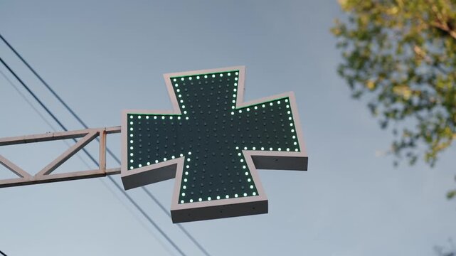 Green pharmacy cross sign with flashing led lights against a clear blue sky, signaling a drugstore with a tree in the background. The lights animate in various patterns to attract attention
