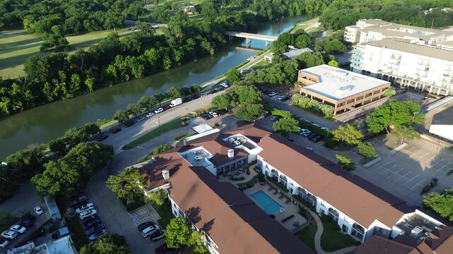 Trinity River curves alongside dense forest belts near residential complexes featuring swimming pools. Brown toned rooftops, green lawns bridge gap to commercial office buildings, Fort Worth, TX