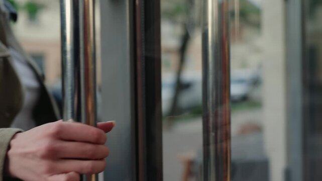 Woman's hand gently touching and pulling a shiny metal handle to open a glass door and enter a building, with a city street reflecting on the surface, showcasing a modern urban lifestyle concept