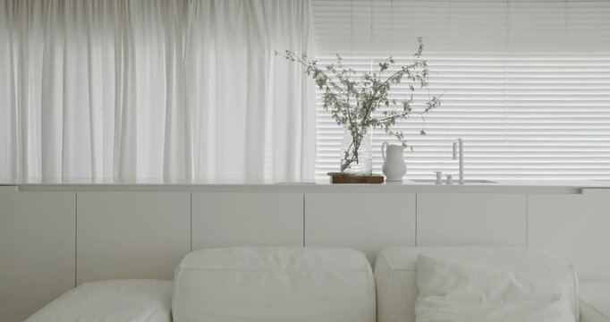 Contemporary white living room or kitchen featuring a modular sofa, delicate branches in a vase, and soft natural light filtering through the blinds and curtains for a serene and elegant ambiance