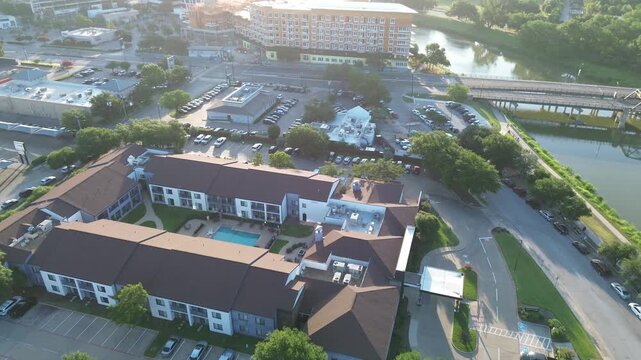 Apartment buildings with brown roofs encircle central swimming pool near Trinity River. Bridges span waterway while wooded buffers separate residential areas from thoroughfares, Fort Worth, TX