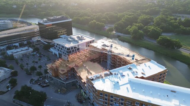 Tower crane looms over building footprint near Trinity River, S University Dr intersection. Golden sunlight illuminates wood skeletons, rooftop membranes, dense tree canopies, Fort Worth, Texas