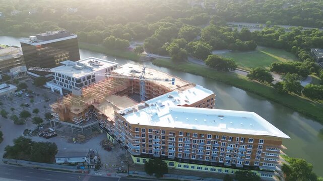 Trinity River winds past emerging multistory structure, bustling S University Dr corridor. Exposed wooden framing contrasts against pristine white roofing, dense greenery, Fort Worth, Texas