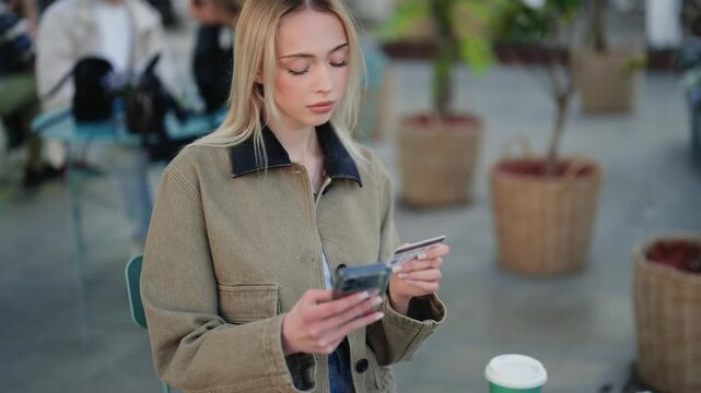 Beautiful blonde woman making an online payment with a credit card and smartphone while sitting at an outdoor cafe. E-commerce and modern technology for convenient digital transactions