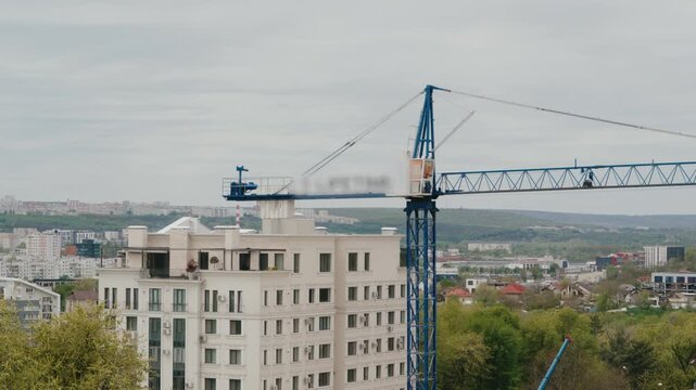 High industrial tower crane rotating its boom over a cityscape with residential buildings and green trees. Construction industry equipment working on a developing urban area during a cloudy day