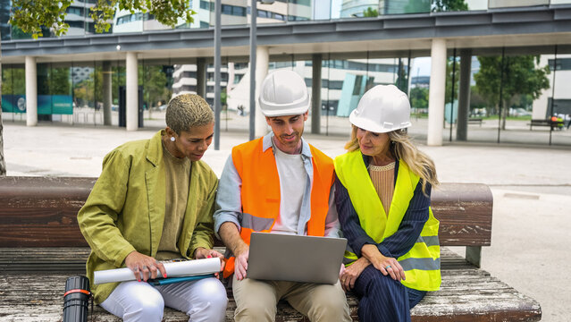 Diverse engineering and architecture professionals discussing project plans and design using a laptop on a city bench