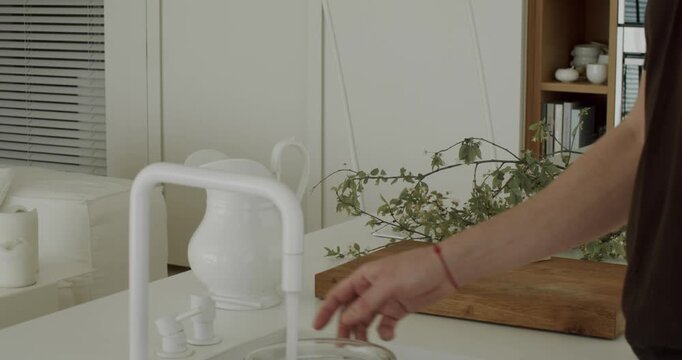 Close up view of a man's hand opening the faucet and washing his hands with running water in a minimalist white kitchen sink, showing a daily routine and hygiene concept