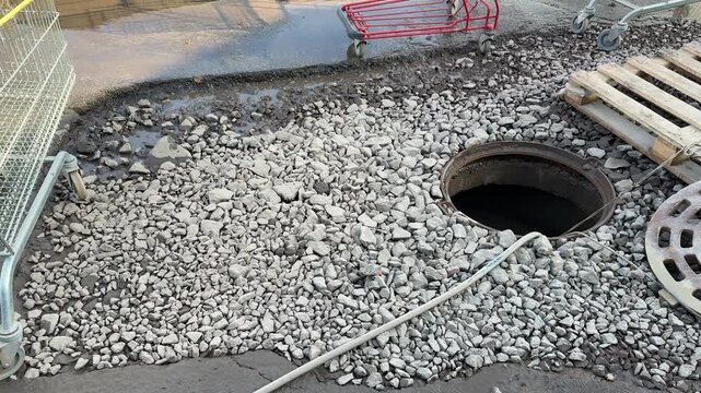 Open inspection well with rubber hose, gravel street, wooden pallet and shopping cart, pump engine running as hose drains water