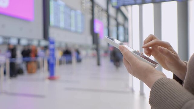 Close-up of hands using a tablet at a modern airport terminal. Background shows blurred passengers and digital signage. Person appears focused and calm while checking travel information