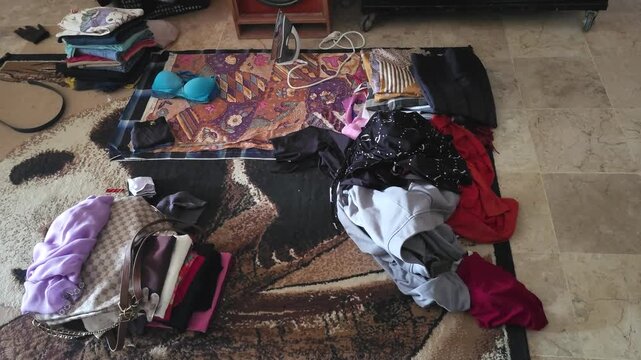 A messy room with scattered clothes on a patterned carpet and tiled floor next to an iron and a handbag