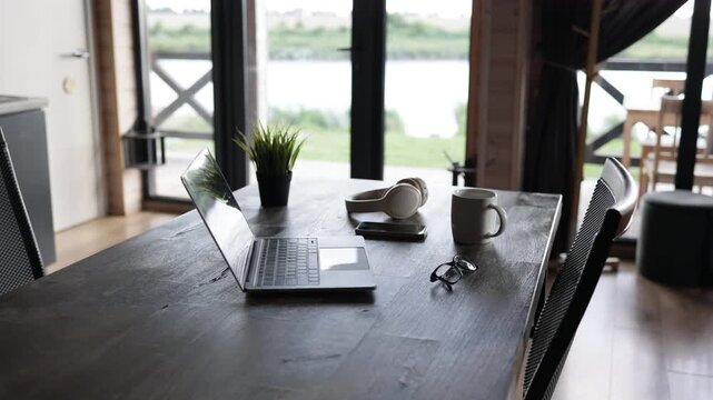 A laptop, headphones, smartphone, and coffee mug are arranged on a rustic wooden table with a scenic view through the window