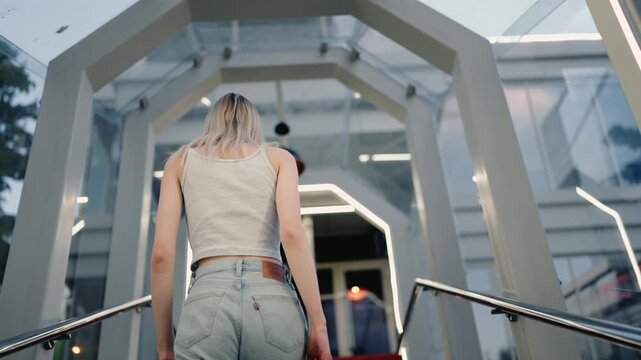 Rear view of a blonde woman in jeans and a tank top walking up stairs in a contemporary architectural setting, showcasing her confident and determined stride as she moves upward