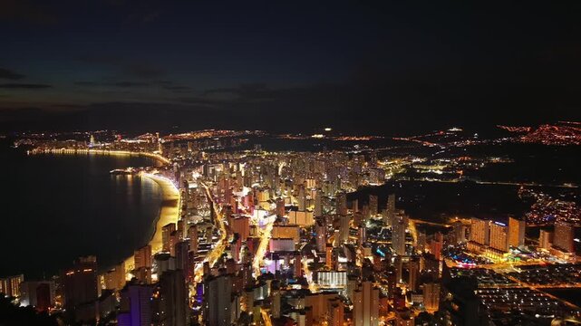Night aerial beach bay illuminated by warm lights and calm water, crescent coastline lit along promenades,