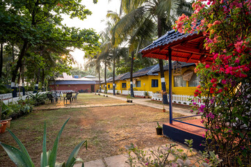 Fototapeta premium Exterior view of yellow cottage houses with blue roofs surrounded by coconut palm trees, illuminated by soft natural light, showcasing charming tropical architecture and a peaceful setting.