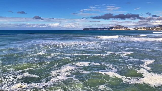 Aerial ocean waves crashing over coastline, turquoise water churning into white foam under bright blue sky,
