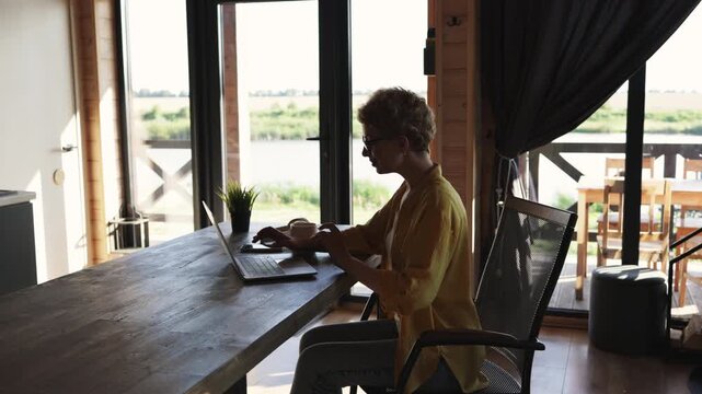 A woman sits at a wooden table, enjoying a cup of coffee while working on her laptop in a bright, modern room with a view of nature