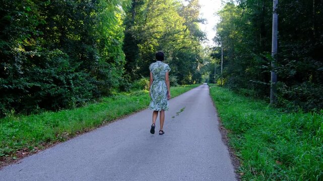 Young woman in green summer dress walks across rustic road . Lush green meadow and forest trees surround narrow dirt path in countryside park.