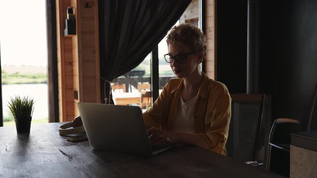 A caucasian woman with short curly hair and glasses holds a mug of coffee while working on her laptop in a rustic cabin setting with a view of a lake
