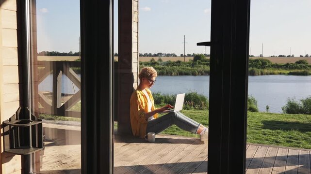 A young caucasian woman with blonde hair sits on a wooden porch, typing on her laptop with a serene lake and green landscape in the background