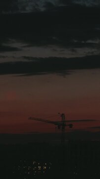 Timelapse of a tall construction crane silhouette against a dramatic, colorful sky during sunset, with dark clouds moving over a city with glowing lights in the background at twilight