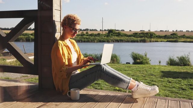 A caucasian woman enjoys a sunny day while working on her laptop by a tranquil lake, embracing a remote work lifestyle