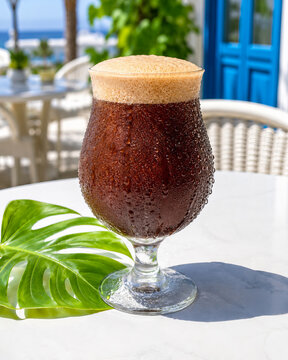 Nitro Cold Brew Coffee in Stemmed Glass with Creamy Foam on White Marble Table, Mediterranean Greek Island Cafe Terrace with Blue Door, Ocean View and Monstera Leaf