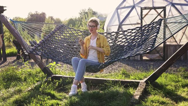 A young caucasian woman wearing headphones and glasses is enjoying music on her phone while relaxing in a hammock in a sunny backyard setting