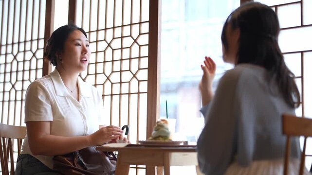 Two women converse over tea in a quiet cafe. They gesture animatedly, sharing thoughts with warmth. Soft light streams through patterned wooden screens. The setting feels calm, intimate