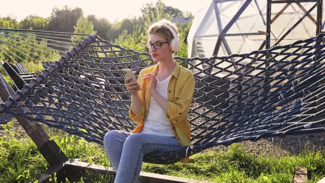 A caucasian woman wearing headphones and glasses sits in a hammock outdoors, holding a smartphone and enjoying music