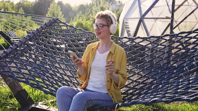 A caucasian woman wearing headphones and glasses is holding a smartphone while relaxing in a hammock outdoors, enjoying music or a podcast