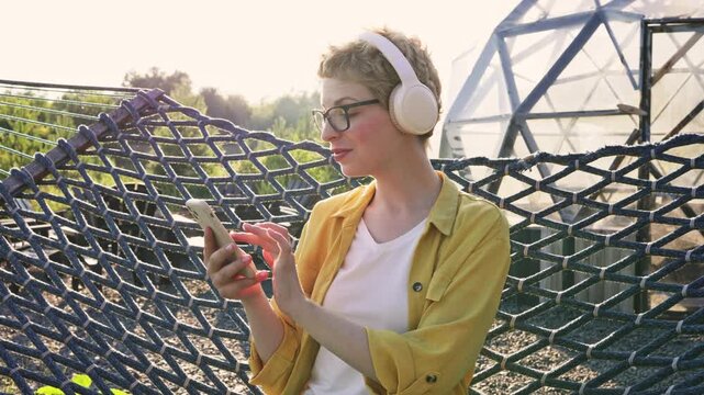 A young woman with short blonde hair and glasses relaxes in a hammock while listening to music on her phone and headphones