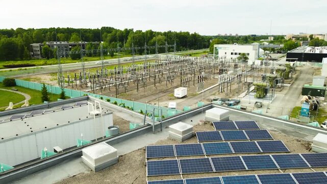 Aerial footage of a high voltage electrical power station with transformers and solar panels on a building rooftop. Showcasing renewable energy generation and electricity distribution infrastructure