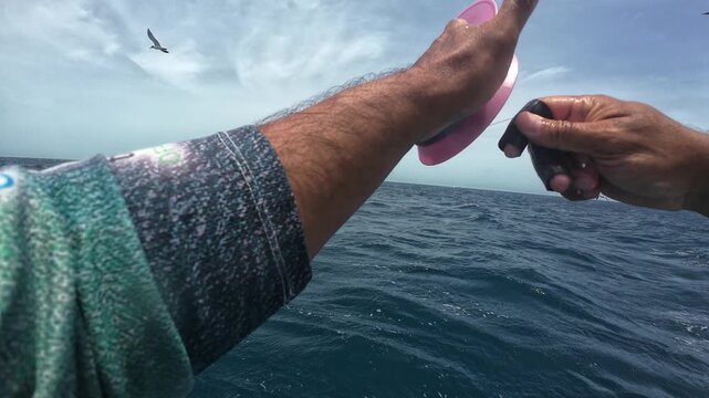 POV first-person perspective of a traditional fisherman manually winding fishing line on a pink spool while seabirds circle in the background.