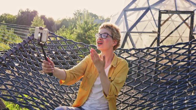 A young woman with short blonde hair and glasses sits in a hammock, holding a selfie stick and blowing a kiss towards the camera