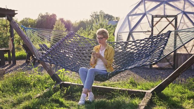 A caucasian woman with short curly hair wearing glasses and casual clothes sits in a hammock outdoors, looking at her smartphone