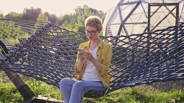 A young caucasian woman with short curly hair and glasses relaxes in a hammock while looking at her smartphone, with a geodesic dome structure in the background