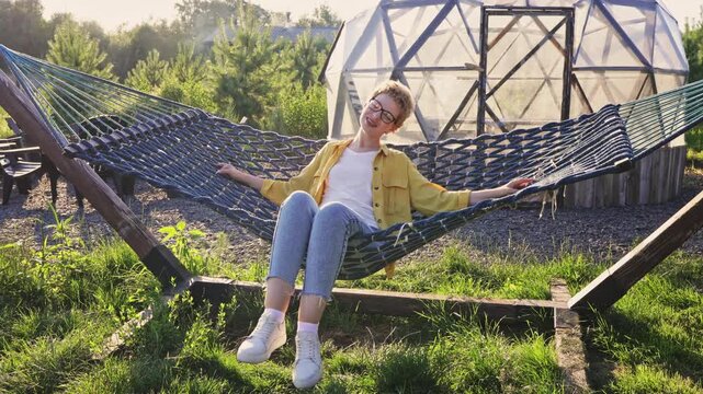 A happy woman with short hair and glasses is relaxing in a hammock, holding a smartphone, with a geodesic dome structure in the background