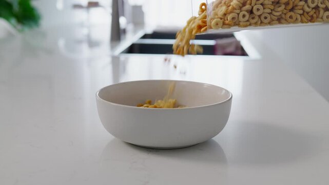 Close-up of breakfast cereal being poured into a bowl on a kitchen benchtop. Crisp flakes cascade into the bowl, capturing a simple morning routine and food preparation scene.