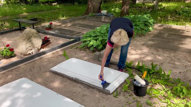 Woman cleaning dust from a restored concrete grave border before painting in a cemetery