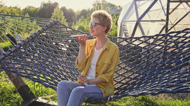 A caucasian woman with glasses sits in a hammock outdoors, holding a phone to her ear and speaking