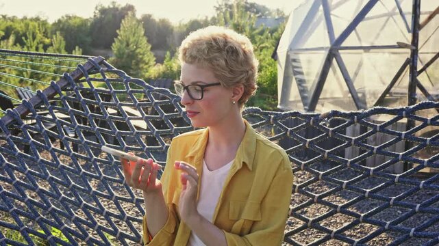 A young caucasian woman with short blonde hair and glasses is sitting on a hammock and talking on her smartphone