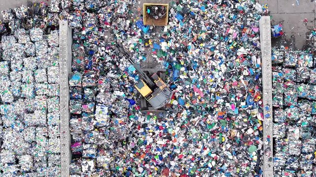 Machines sort and process plastic waste in a recycling facility. Large piles of mixed recyclable materials are visible. Sorting equipment operates to separate different plastics.