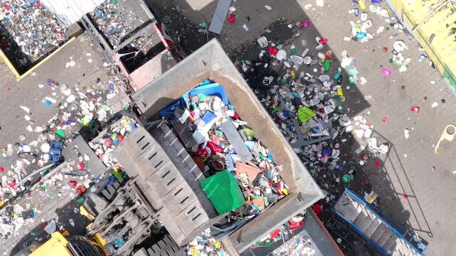 Trucks unload plastic waste at a recycling facility. Workers sort materials for processing. Machinery handles plastic for recovery and sorting. Waste management operations support recycling.