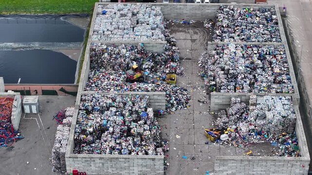 A large waste recycling facility processes plastic waste. Containers hold sorted plastic materials. Workers manage operations at the industrial site. Various recyclable items are visible.