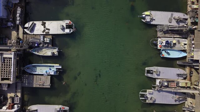 Drone shot of traditional fishing and oyster barges moored along the narrow water canals of Port Leucate, Occitanie, France. Crystal clear green water with visible seabed.
