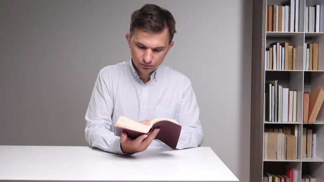 Man focusing on a book while sitting at a white desk with a bookshelf filled with various titles in the background, representing education, knowledge, and personal development