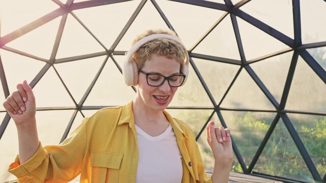 A young Caucasian woman with short blonde hair and glasses enjoys music while dancing inside a geodesic dome structure